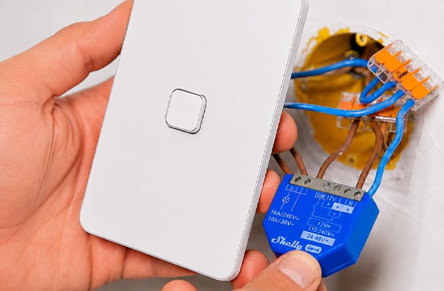 A hand holding a white light switch plate away from the wall, with a smart module wired into the box behind it
