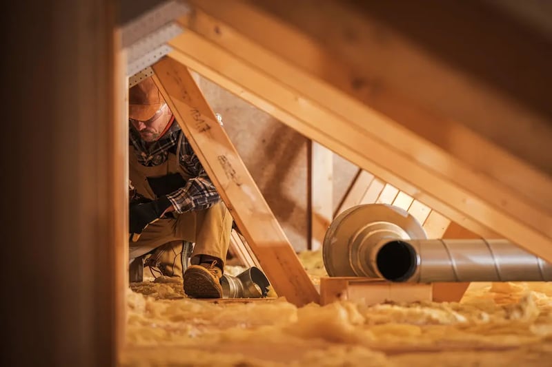 A tradesperson kneeling among the trusses in a roof space, beside an exhaust fan and its silver duct lying on the insulation.
