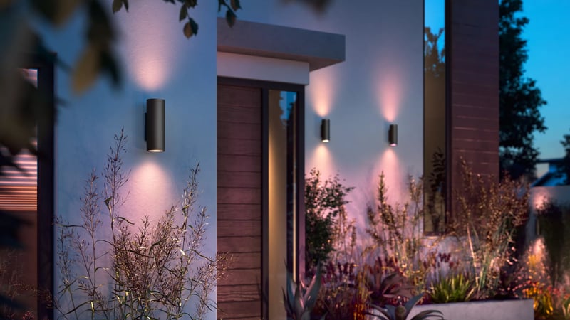 The front of a house at dusk, with wall lights glowing either side of the front door and a garden bed in front