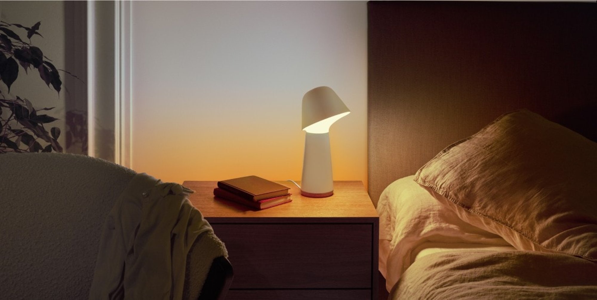 A bedside table with a white lamp and two books, the wall behind glowing sunrise orange beside an unmade bed.