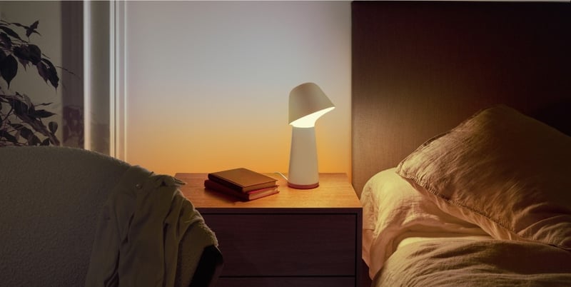 A bedside table with a white lamp and two books, the wall behind glowing sunrise orange beside an unmade bed.