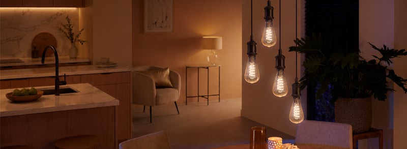 An open kitchen and lounge in the evening, with pendant bulbs over the table, a strip light under the cabinets and a table lamp all on.