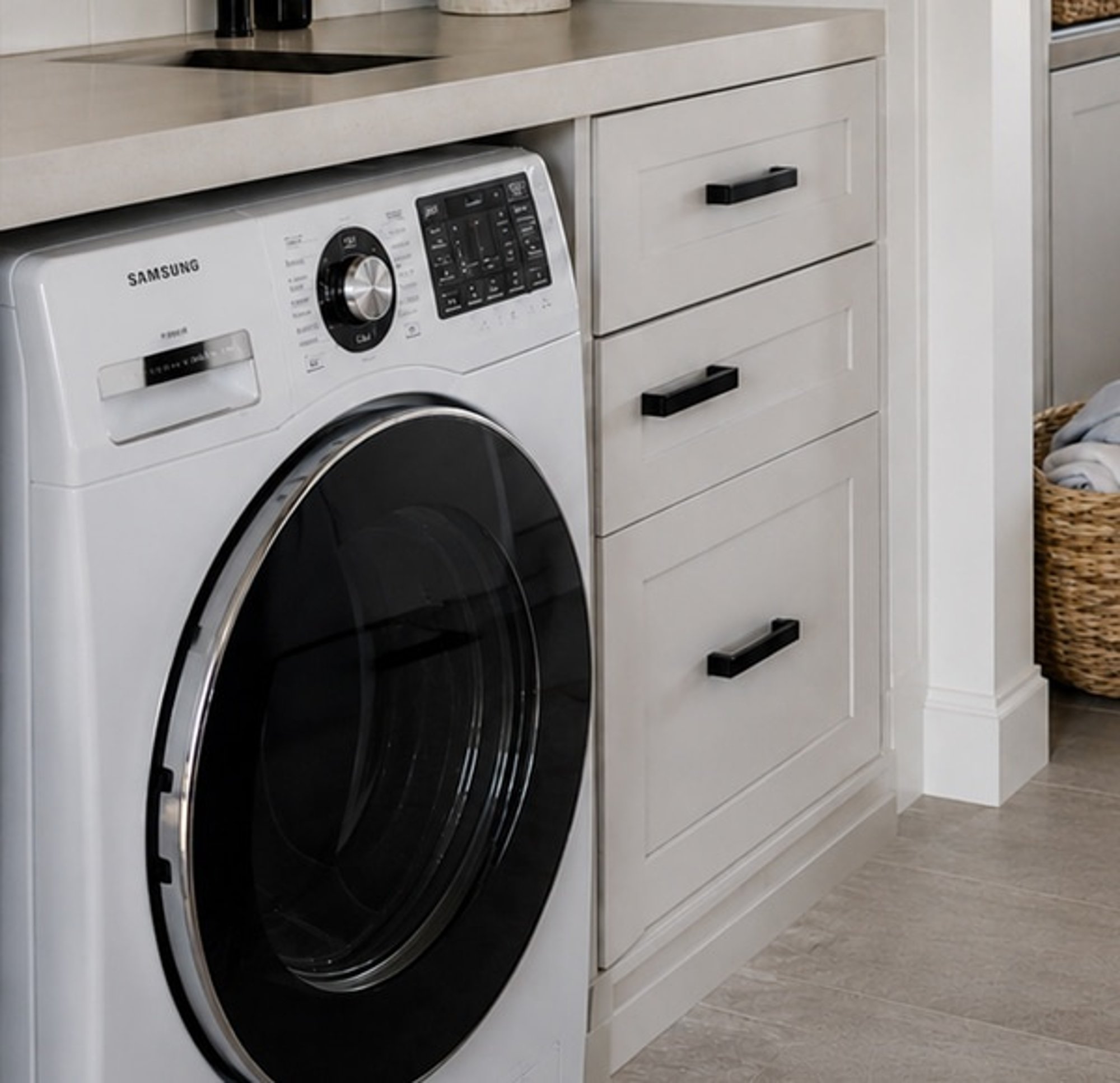 A front-loading washing machine under the bench in a laundry, beside a bank of drawers