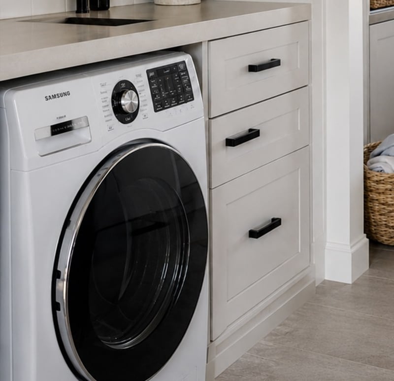 A front-loading washing machine under the bench in a laundry, beside a bank of drawers