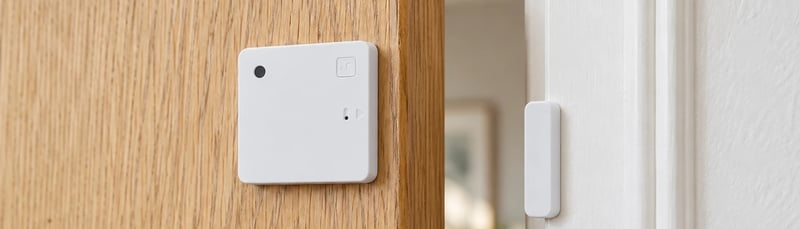 A white Shelly BLU Door/Window sensor on the edge of an oak door, with its magnet on the white door frame beside it